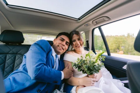 Hombre con traje azul y mujer con vestido blanco y ramo están sentados juntos dentro de un automóvil.