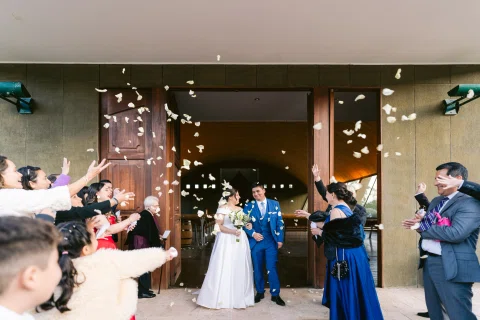 Hombre con traje azul y mujer con vestido blanco salen por una puerta mientras varias personas lanzan pétalos.