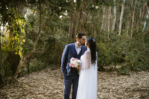 Hombre con traje azul y mujer con vestido blanco se besan entre árboles.