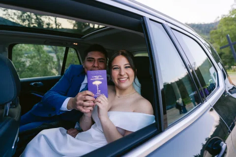 Hombre con traje azul y mujer con vestido blanco sonríen desde un automóvil mientras muestran un cuadernillo morado.