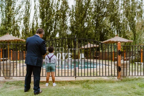 Hombre con traje azul y niño con suspensores de pie junto al cerco de una piscina.