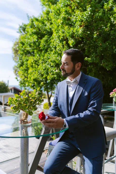 Hombre con traje azul sentado al aire libre junto a una mesa de vidrio.