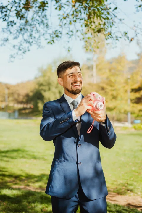 Hombre con traje azul sonríe mientras sostiene una cámara instantánea rosada en el parque.