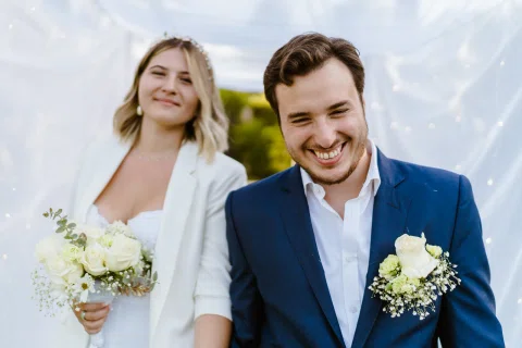 Hombre con traje azul sonríe hacia la cámara mientras una mujer vestida de blanco aparece detrás con un ramo.