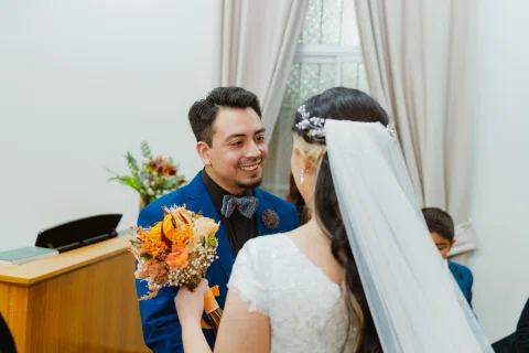 Hombre con traje azul sonríe frente a una mujer con vestido blanco que sostiene un ramo de flores naranjas.