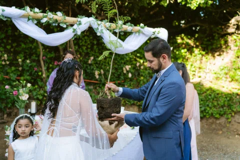 Hombre con traje azul sostiene un árbol joven con el cepellón de tierra visible frente a una mujer con vestido blanco.