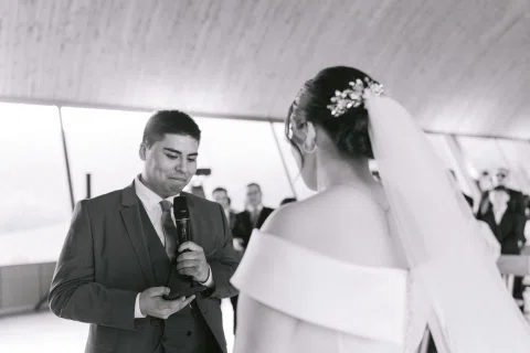 Hombre con traje azul sostiene un micrófono y un objeto mientras una mujer con vestido blanco lo observa, en blanco y negro.