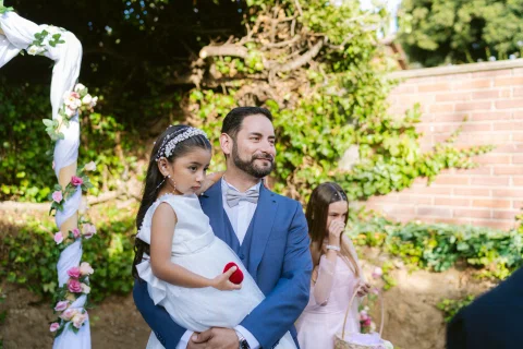 Hombre con traje azul sostiene en brazos a una niña con vestido blanco; otra niña aparece detrás.