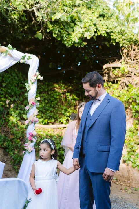Hombre con traje azul toma de la mano a una niña con vestido blanco junto a un arco floral.