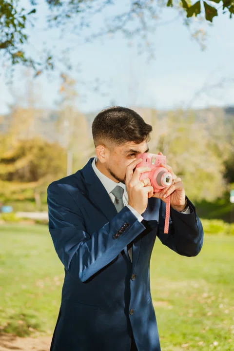 Hombre con traje azul usa una cámara instantánea rosada al aire libre.