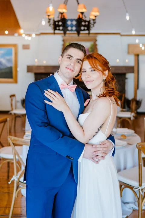 Hombre con traje azul y mujer con vestido blanco permanecen abrazados dentro de un salón.