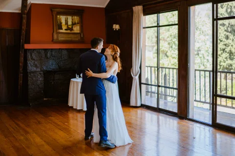Hombre con traje azul y mujer con vestido blanco permanecen abrazados dentro del salón junto a grandes ventanales.