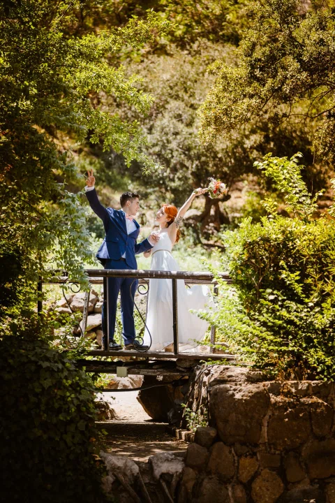 Hombre con traje azul y mujer con vestido blanco levantan los brazos sobre un pequeño puente rodeado de vegetación.