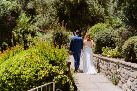 Hombre con traje azul y mujer con vestido blanco caminan de espaldas por un puente de piedra en el jardín.