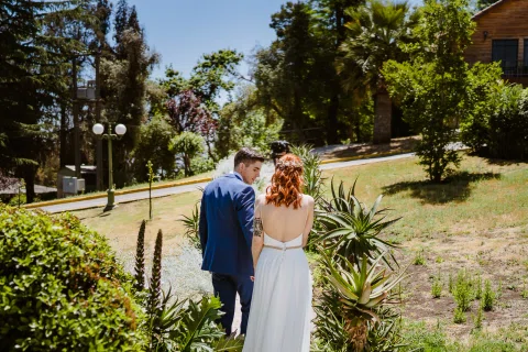 Hombre con traje azul y mujer con vestido blanco caminan de espaldas por un sendero entre plantas del jardín.