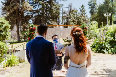 Hombre con traje azul y mujer con vestido blanco vistos de espaldas frente a una persona que sostiene documentos en un jardín.