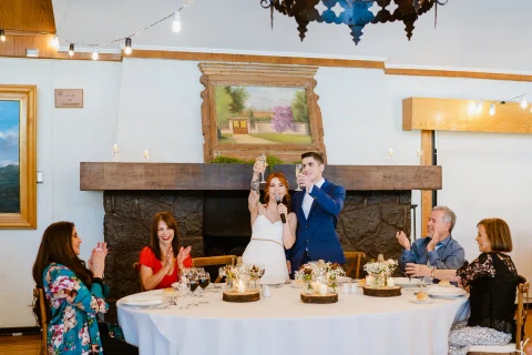 Hombre con traje azul y mujer con vestido blanco permanecen de pie junto a una mesa mientras varias personas están sentadas alrededor.