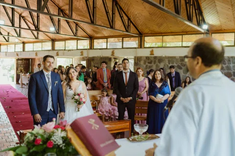 Hombre con traje azul y mujer con vestido blanco permanecen frente a un hombre que sostiene un libro dentro de la iglesia.