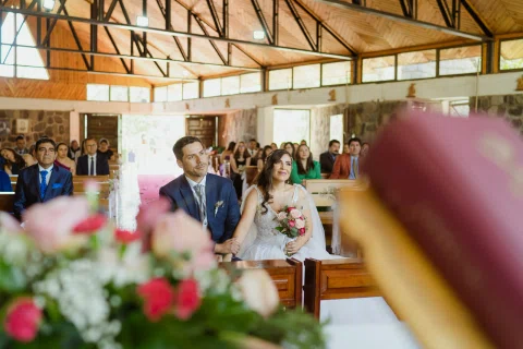 Hombre con traje azul y mujer con vestido blanco permanecen juntos en la zona del altar, con flores desenfocadas en primer plano.