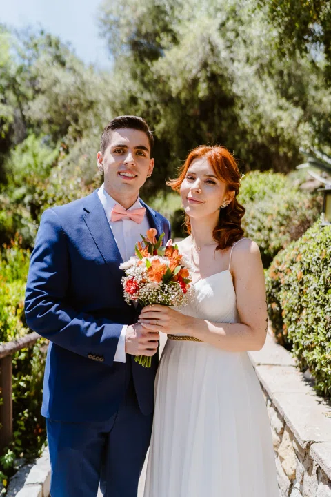Hombre con traje azul y mujer con vestido blanco posan juntos sosteniendo un ramo en el jardín.