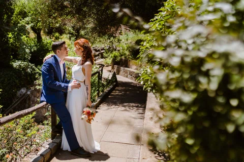 Hombre con traje azul y mujer con vestido blanco posan juntos junto a una baranda entre vegetación.