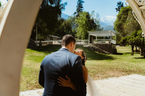 Hombre con traje azul y mujer con vestido blanco se besan en un jardín con una montaña al fondo.