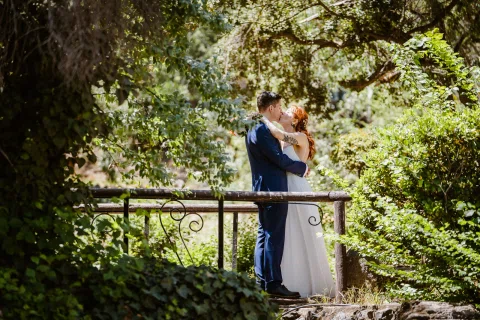 Hombre con traje azul y mujer con vestido blanco se besan sobre un pequeño puente rodeado de vegetación.