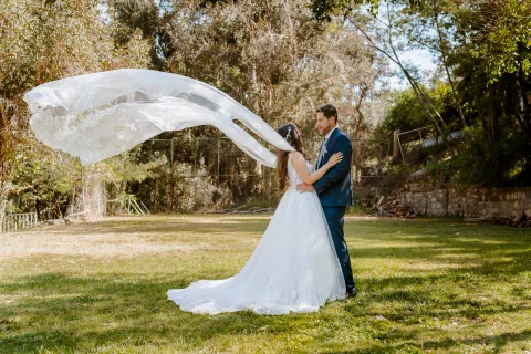 Hombre con traje azul y mujer con vestido blanco posan en un jardín mientras el velo de ella se extiende en el aire.