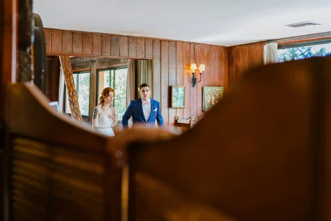 Hombre con traje azul y mujer con vestido blanco visibles al fondo entre una estructura de madera dentro del salón.