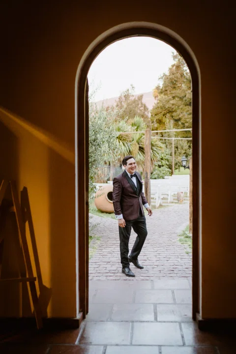 Hombre con traje burdeos camina hacia una puerta, visto a través de una abertura arqueada desde un interior oscuro.