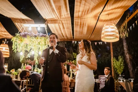 Hombre con traje burdeos habla con un micrófono mientras una mujer con vestido blanco permanece a su lado en la recepción.