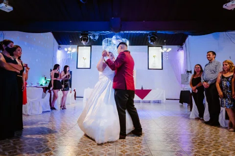Hombre con traje burdeos y mujer con vestido blanco bailan en un salón iluminado, rodeados por otras personas.