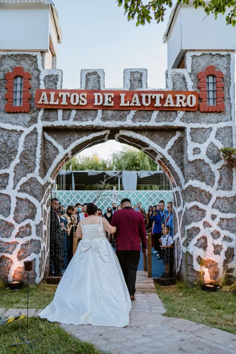 Hombre con traje burdeos y mujer con vestido blanco vistos de espaldas bajo un arco de piedra, con varias personas al otro lado.