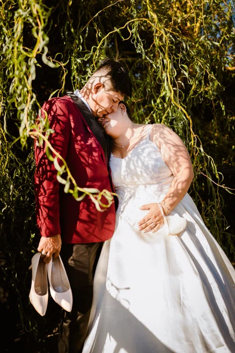 Hombre con traje burdeos y mujer con vestido blanco juntan sus cabezas junto a ramas colgantes; él sostiene un par de zapatos.