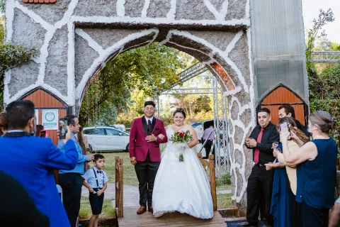 Hombre con traje burdeos y mujer con vestido blanco caminan bajo un arco de piedra en el exterior.