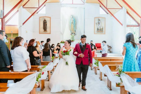 Hombre con traje burdeos y mujer con vestido blanco caminan por el pasillo de una iglesia entre personas de pie.