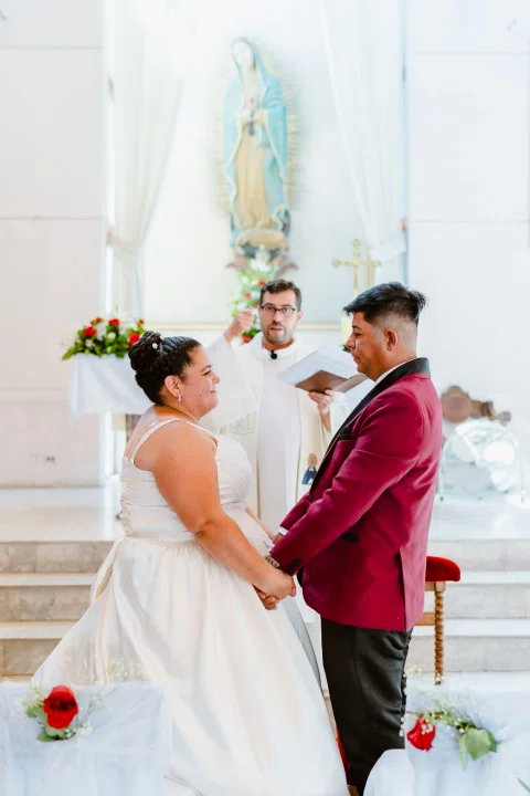 Hombre con traje burdeos y mujer con vestido blanco de pie frente a una persona con vestimenta blanca.