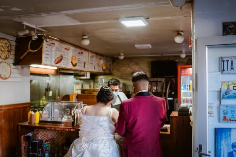 Hombre con traje burdeos y mujer con vestido blanco están de pie frente al mostrador de un local de comida.