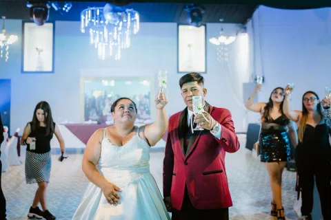 Hombre con traje burdeos y mujer con vestido blanco levantan copas mientras posan en una pista de baile.