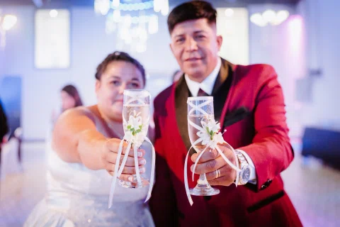 Hombre con traje burdeos y mujer con vestido blanco muestran hacia la cámara dos copas decoradas.
