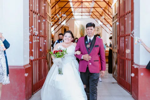 Hombre con traje burdeos y mujer con vestido blanco salen juntos por una puerta roja.