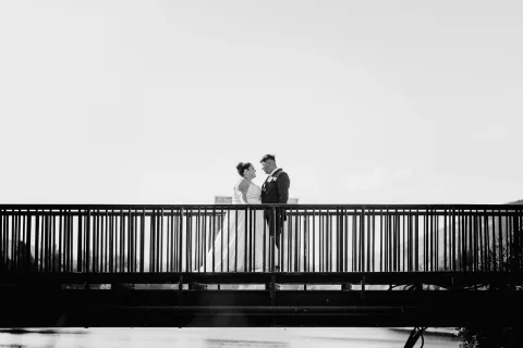 Hombre con traje burdeos y mujer con vestido blanco se besan sobre un puente en una fotografía en blanco y negro.