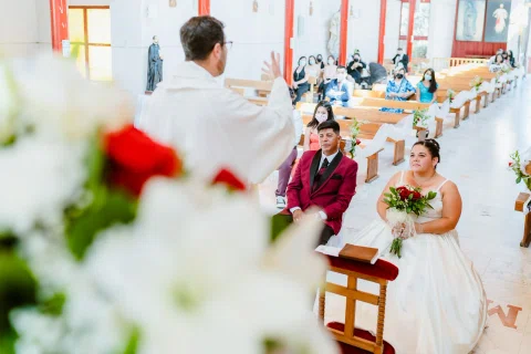 Hombre con traje burdeos y mujer con vestido blanco sentados, vistos detrás de flores desenfocadas.