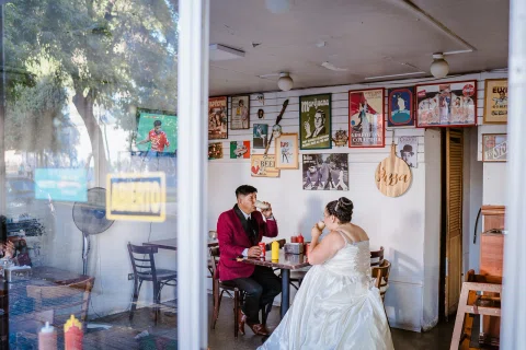 Hombre con traje burdeos y mujer con vestido blanco están sentados frente a frente en una mesa dentro de un local, vistos a través de una ventana.