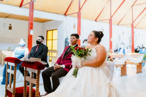 Hombre con traje burdeos y mujer con vestido blanco sentados, con otras personas al fondo.
