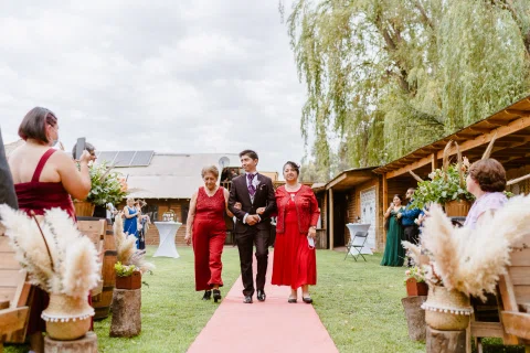 Hombre con traje oscuro camina por la alfombra roja tomado del brazo de dos mujeres vestidas de rojo.