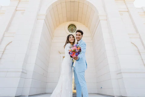 Hombre con traje celeste abraza a una mujer con vestido blanco bajo un gran arco del edificio.
