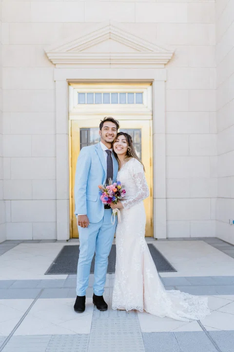 Hombre con traje celeste y mujer con vestido blanco se abrazan frente a una puerta dorada.
