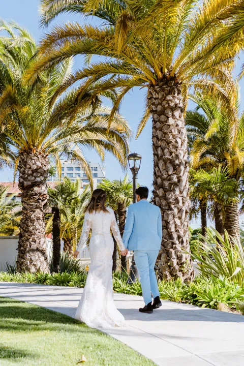 Hombre con traje celeste y mujer con vestido blanco caminan de espaldas por un sendero entre palmeras.