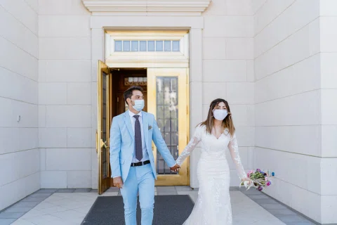 Hombre con traje celeste y mujer con vestido blanco caminan tomados de la mano frente a un edificio blanco.